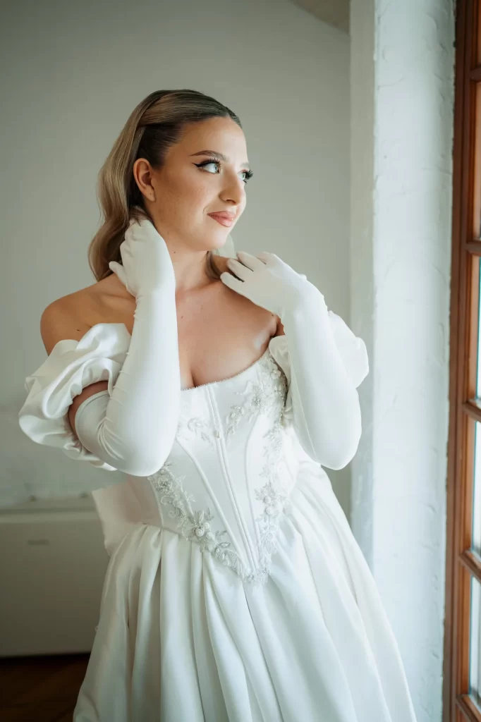 Miruna & Ionuț — Portret al miresei la fereastră cu lumină laterală și gest elegant.