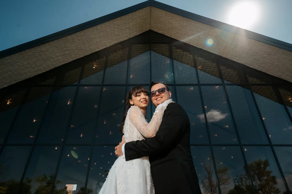 Raluca & Marius — Îmbrățișare cu zâmbet și fațada locației în fundal.